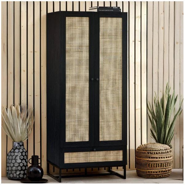 Elegant black Padstow wardrobe standing against a slatted wooden wall, highlighting woven rattan panels and metal base.