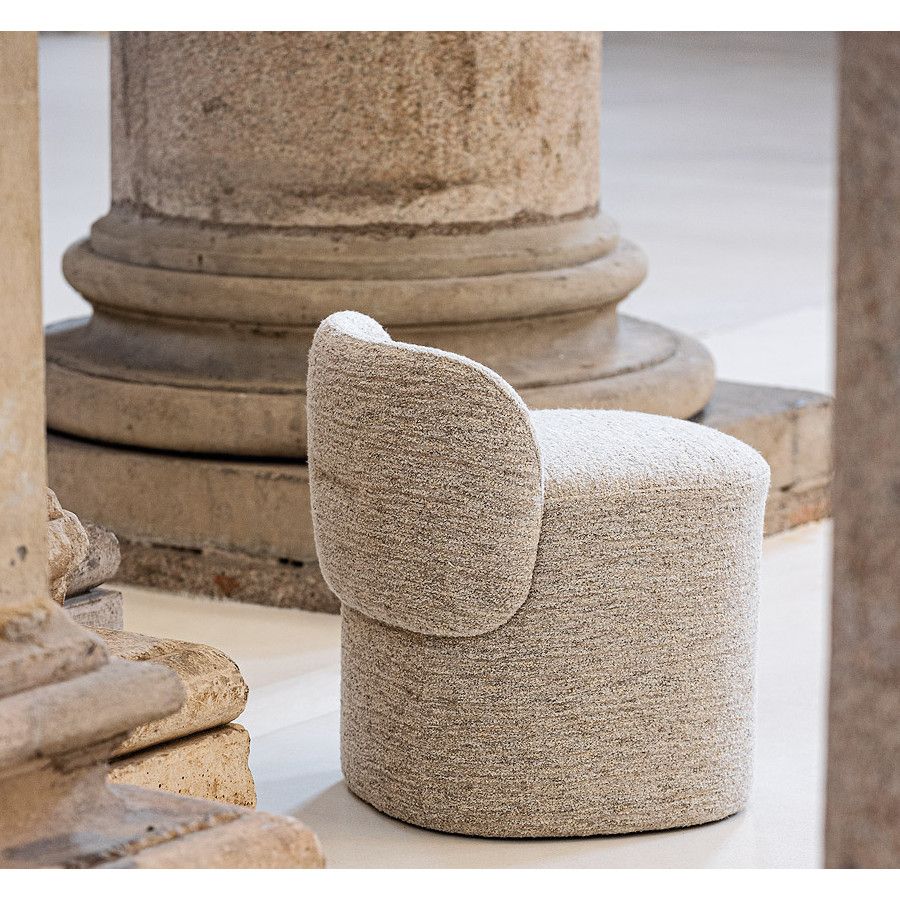Amina Pouf in textured beige upholstery, rear-side profile showing rounded drum base and curved low backrest beside stone columns